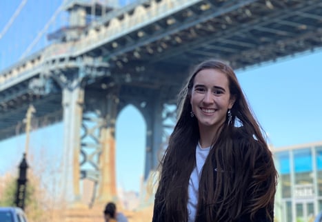 The author, a woman in her 20s with waist-length brown hair, smiling in front of the Brooklyn Bridge.