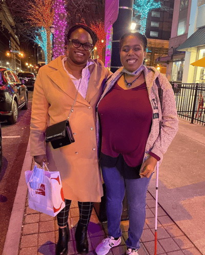 Lily Mourdant, smiling in a jacket, magenta shirt and wearing a necklace. She is standing next to her friend who is wearing a beige trench coat and smiling.