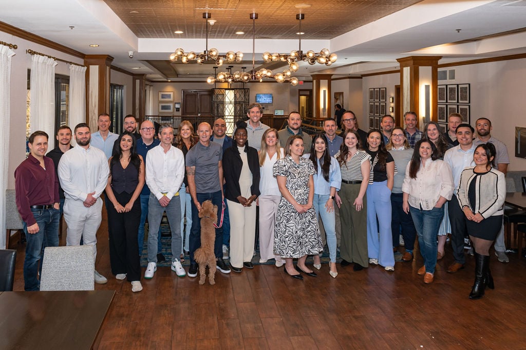 Group photo of the UsableNet U.S. sales team gathered indoors in Austin, Texas, celebrating the company’s 25th anniversary. Approximately 35 people are smiling at the camera, standing in rows, with one small brown dog in the front. The setting features warm lighting, wood floors, and modern decor.