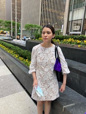 Photo of Emely standing outside in a city square. She has fair skin, dark brown hair and is wearing a white lace dress with a purple handbag on her shoulder.